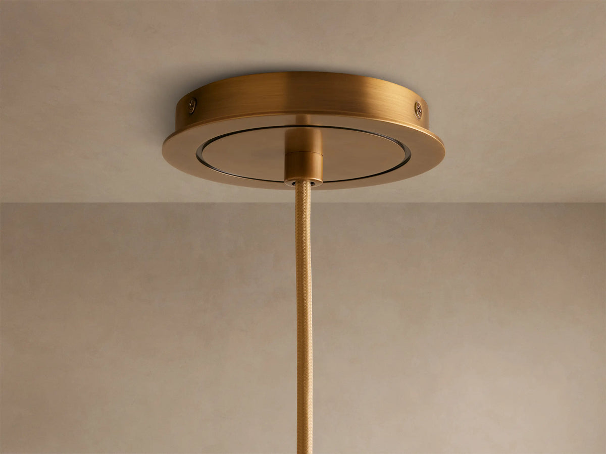 A cylindrical brass pendant light with a smooth, brushed finish, hanging downward against a neutral beige background, illuminated warmly.