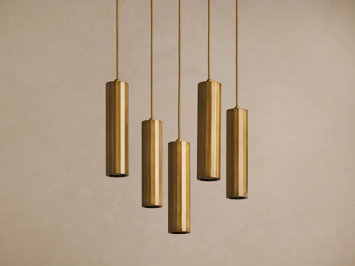 Five cylindrical pendant lights in a brass finish hang vertically against a plain beige background.