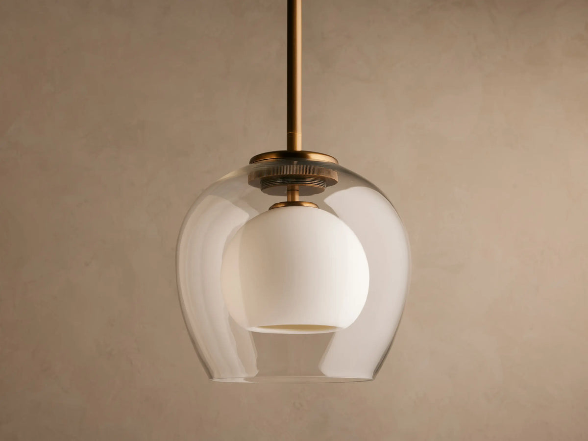 A modern pendant light with a clear glass dome and inner white globe, suspended from a brass rod, illuminated against a beige wall.