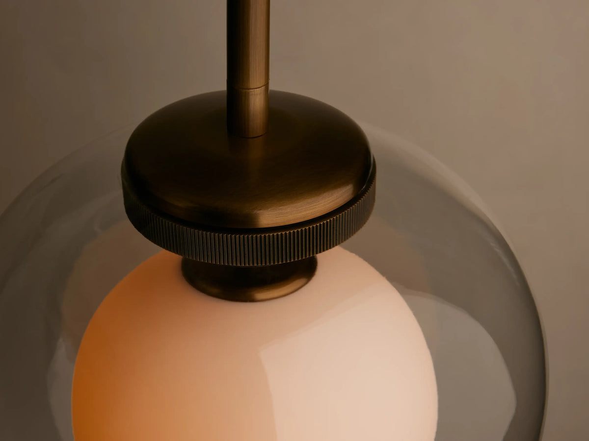 A pendant light with a clear glass dome and an inner white frosted shade, suspended from a brass rod against a beige background.