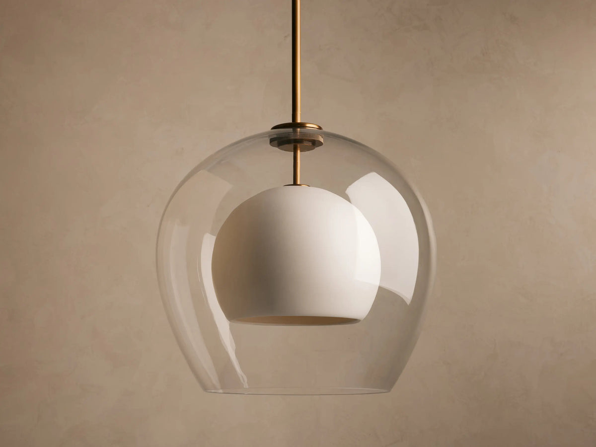 A glass pendant light with an inner frosted globe, suspended from a brass rod against a neutral wall, softly illuminated.