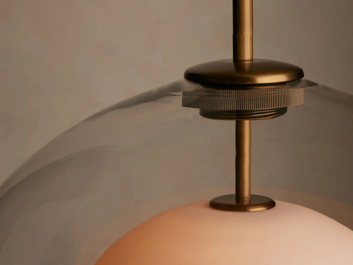 Hanging pendant light with a transparent glass dome and white inner sphere, suspended from a brass rod against a beige background.