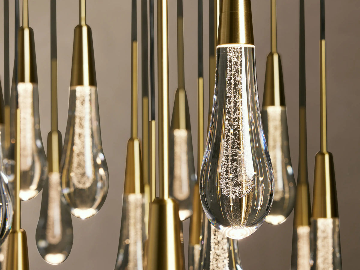 Teardrop-shaped glass bulbs hang from a brass-finished steel Soltaire Round Chandelier, showcasing integrated LED lights.