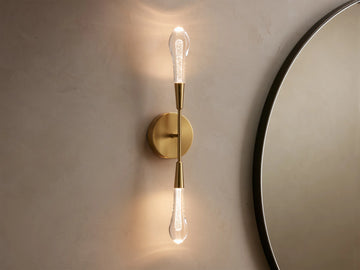 Soltaire Double Sconce with a brass finish, two elongated LED bulbs vertically aligned on a circular backplate against a beige wall.