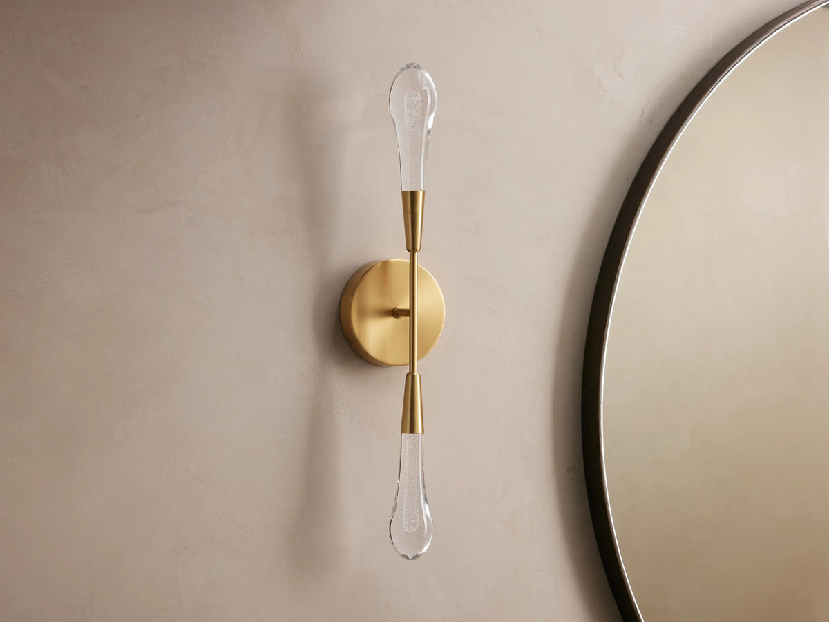 Soltaire Double Sconce with a brass finish, two elongated LED bulbs vertically aligned on a circular backplate against a beige wall.