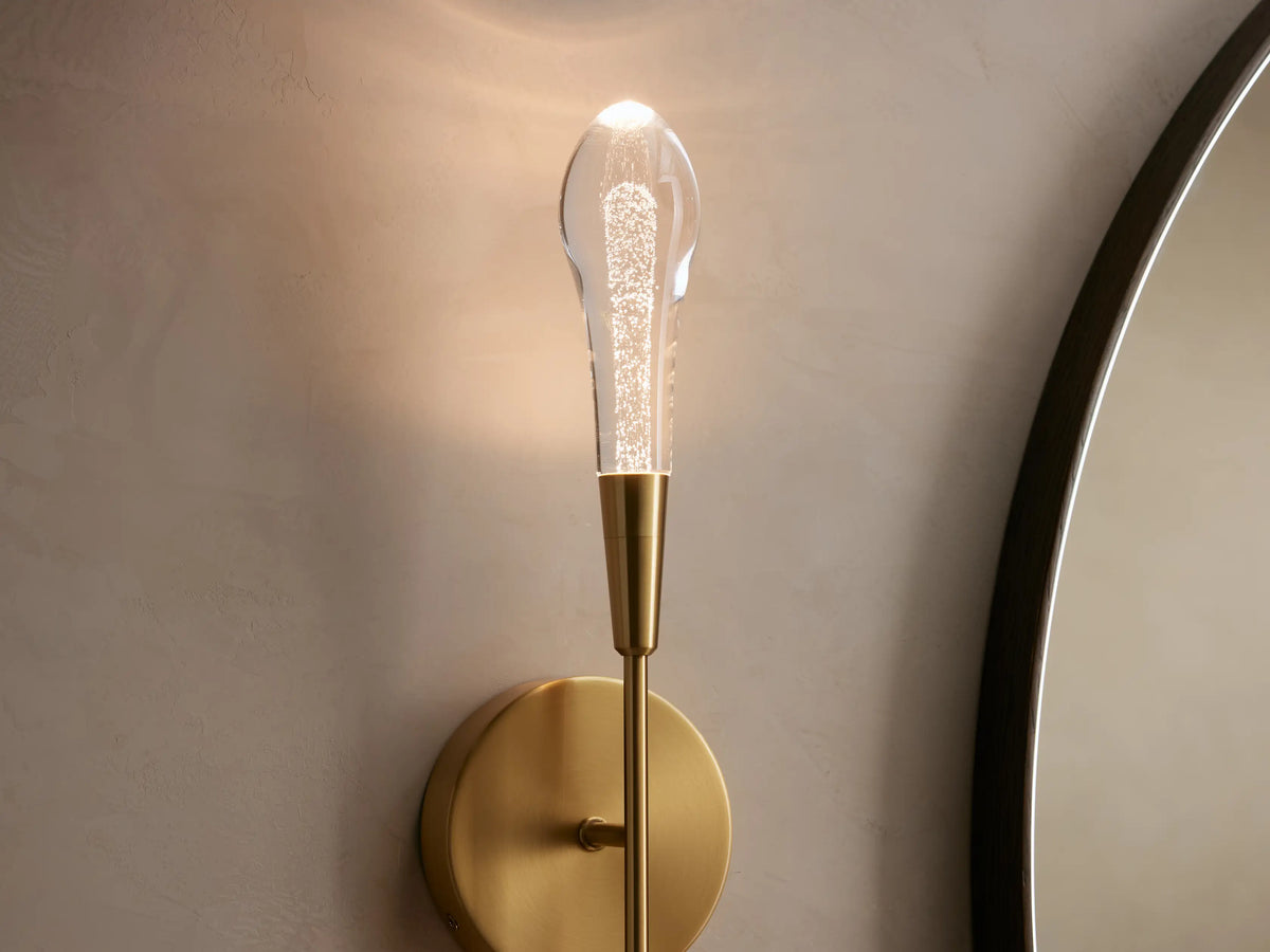 Soltaire Double Sconce with brass finish, featuring two clear elongated bulbs, mounted on a round wall plate against a neutral wall.