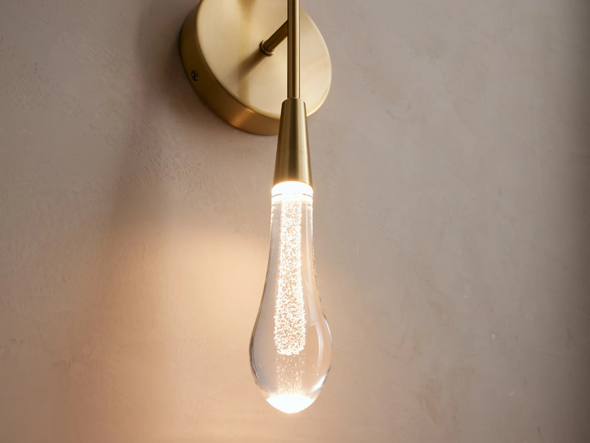 Solitaire Double Sconce with brass finish mounted on a wall, features a clear bulb emitting light, showcasing its elegant design.