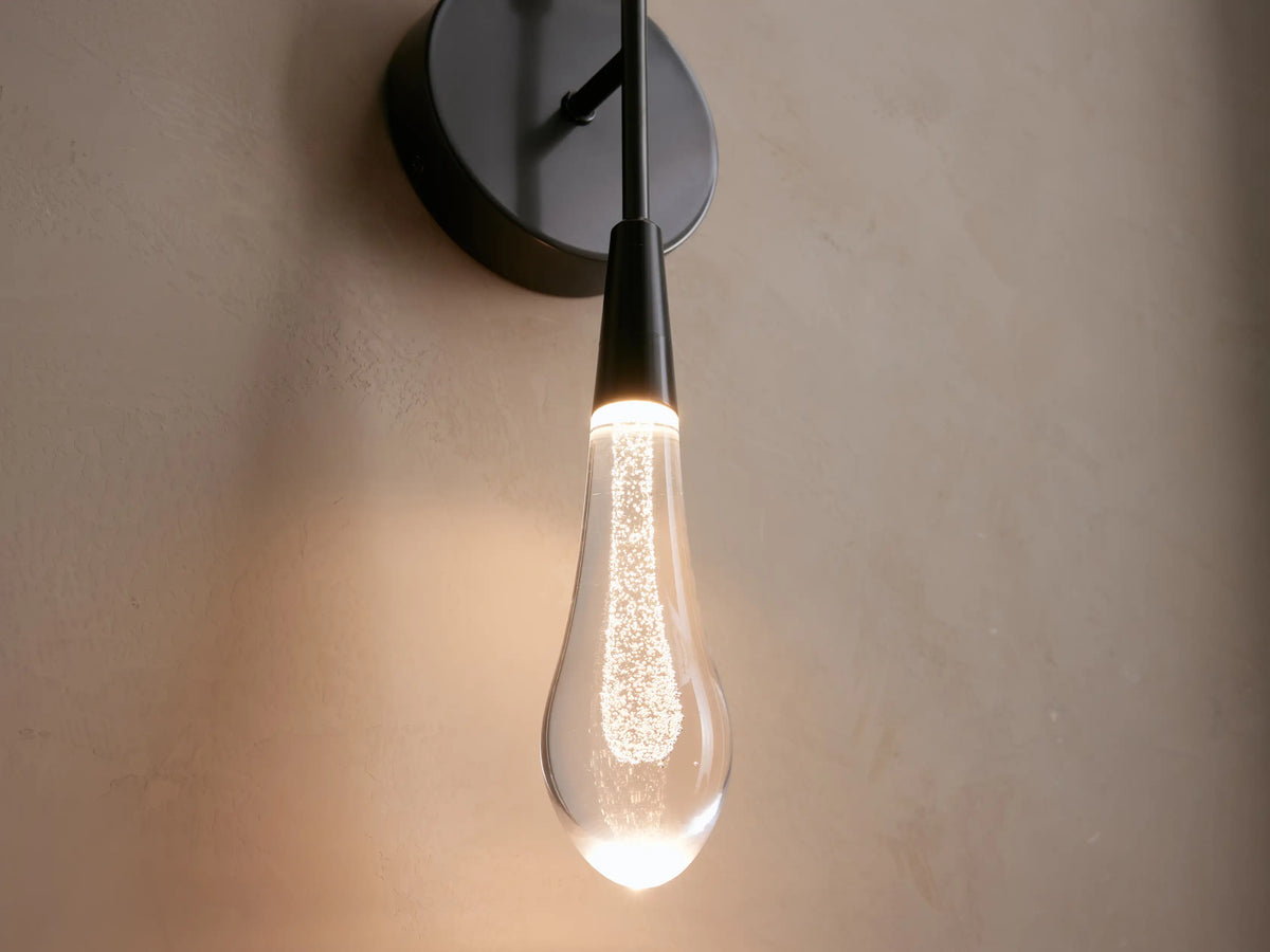 Soltaire Double Sconce with a sleek black backplate and elongated clear glass bulb, mounted on a light-colored wall, partially lit.