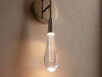 Soltaire Double Sconce with a nickel finish, featuring a teardrop-shaped glass bulb glowing warmly, mounted on a round backplate against a beige wall.
