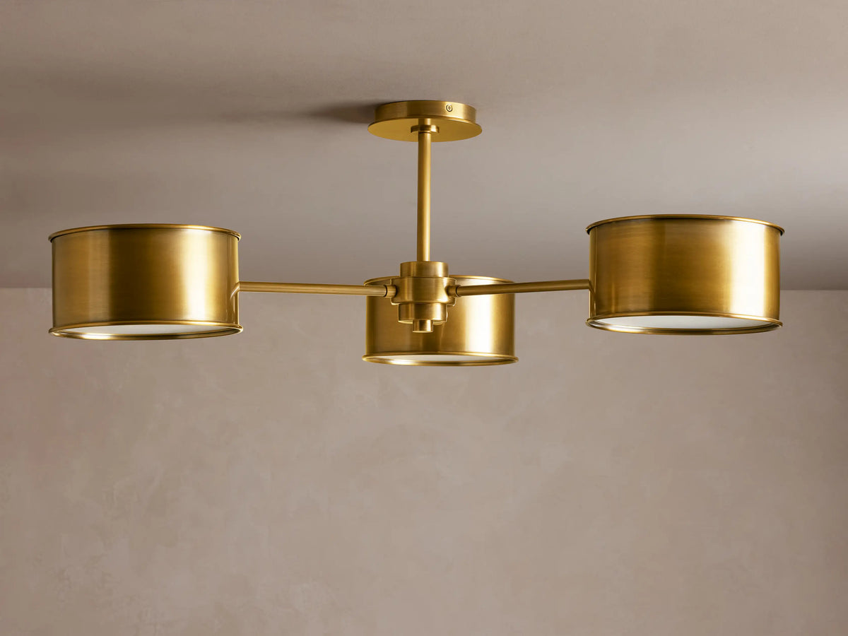 Daphne Metal Flush Mount with three antique brass cylindrical shades, glowing warmly, mounted on a light ceiling.