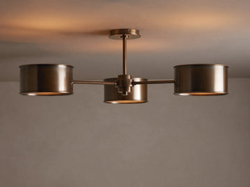 Daphne Metal Flush Mount in warm bronze with three cylindrical shades casting a soft, warm glow on a light ceiling.