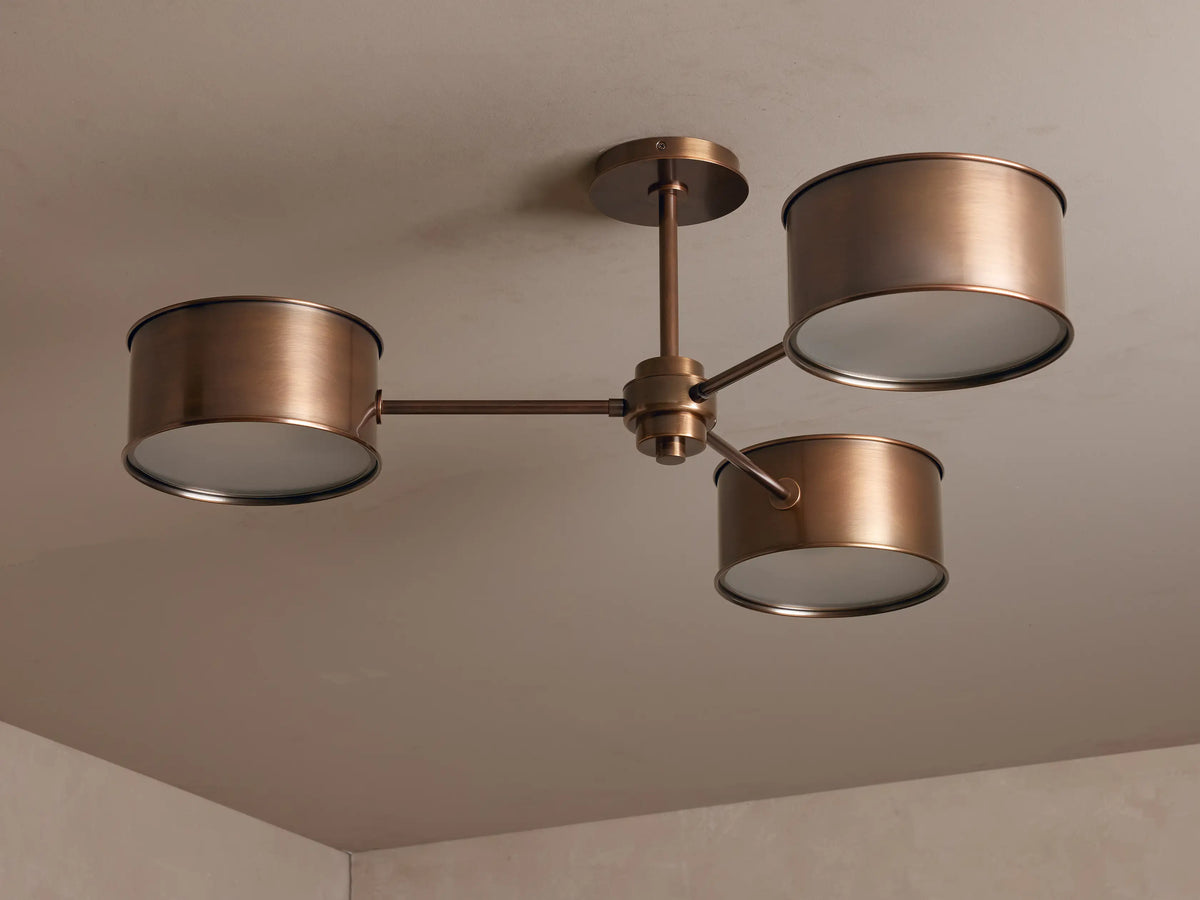Daphne Metal Flush Mount: 3-light antique brass ceiling fixture with cylindrical shades emitting a warm glow on a light ceiling.