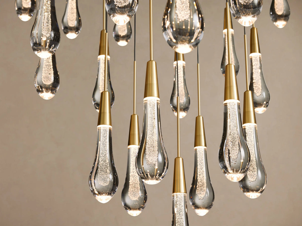 Close-up of Soltaire Round Chandelier, showing multiple brass-capped, bubbly clear glass teardrop pendants glowing.