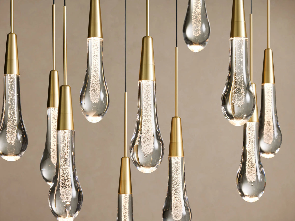 Soltaire Linear Chandelier with multiple glowing, teardrop-shaped clear glass pendants filled with bubbles, suspended at varying heights from brass rods.