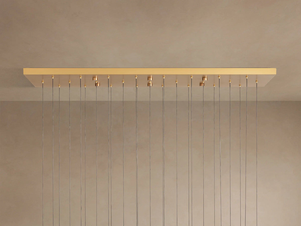 Soltaire Linear Chandelier with a rectangular brass-finished canopy mounted on a light ceiling, from which many thin wires hang.