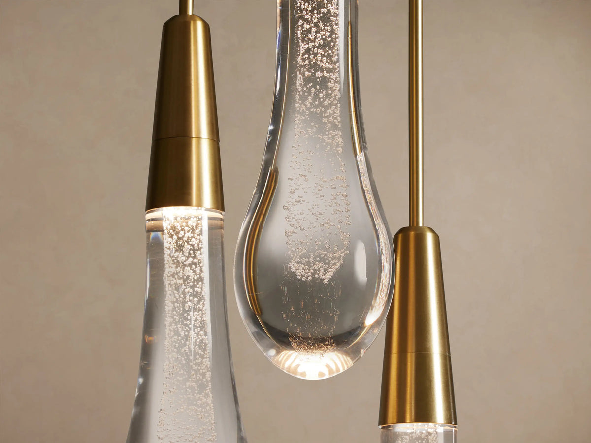 Close-up of Soltaire Linear Chandelier showing three pendants with brass conical tops and glowing, bubble-filled glass teardrops.
