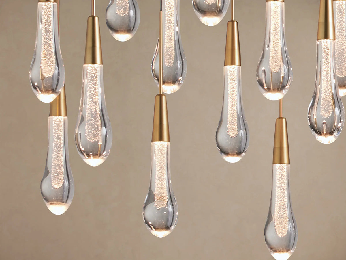 Close-up of Soltaire Linear Chandelier. Many clear glass teardrop pendants with internal bubbles & brass caps emit warm light.