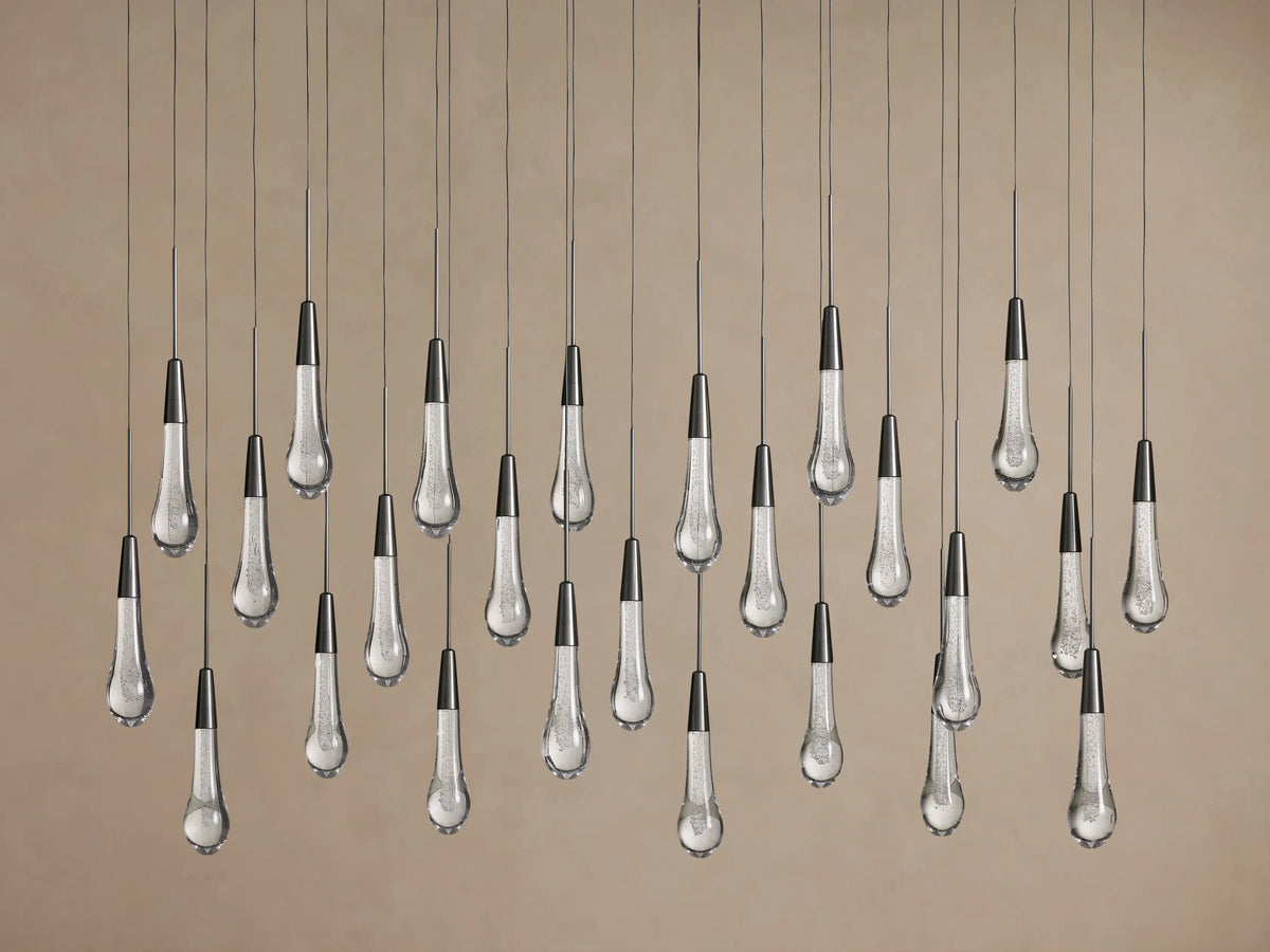 Soltaire Linear Chandelier: Many clear glass teardrop pendants with bubbles & dark metal tops hang against a beige wall.