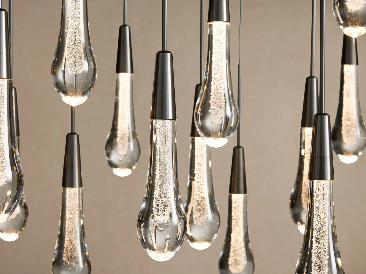 Soltaire Linear Chandelier: Many glowing clear glass teardrop pendants with internal bubbles, suspended from dark metal caps.