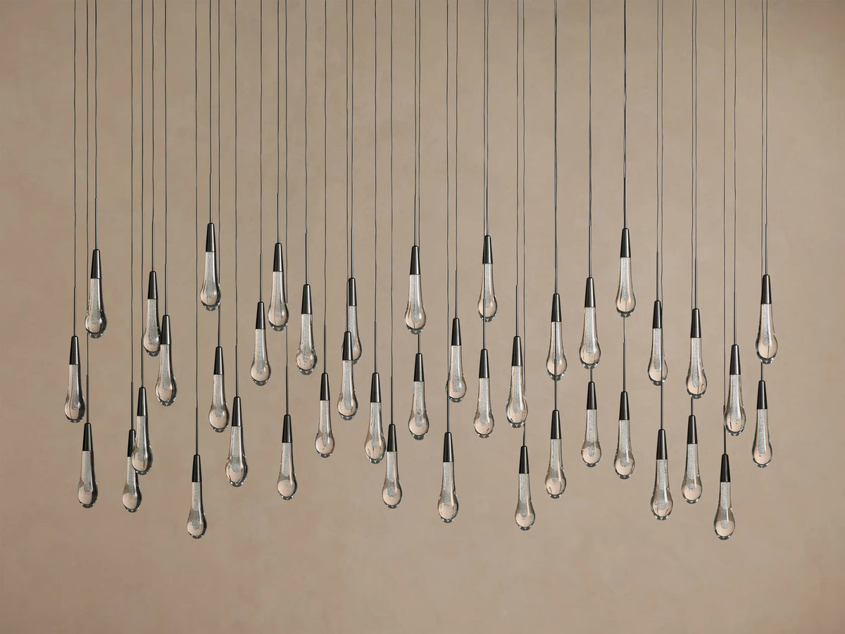 Soltaire Linear Chandelier features multiple clear glass teardrop pendants with dark tops, suspended at varied heights on beige.