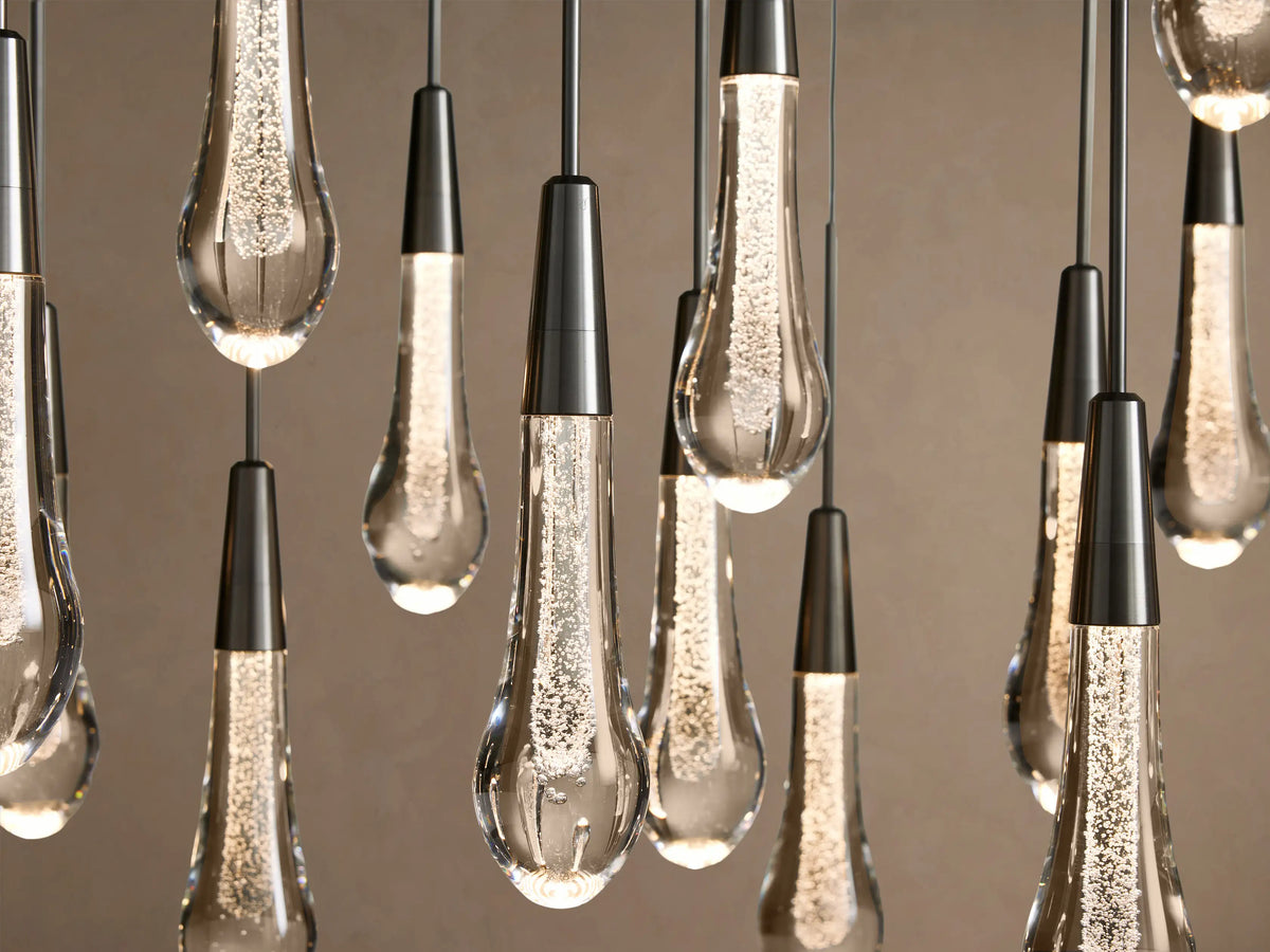 Close-up of Soltaire Linear Chandelier with multiple clear teardrop glass pendants, each glowing brightly with internal bubbles, hanging from dark metal caps.