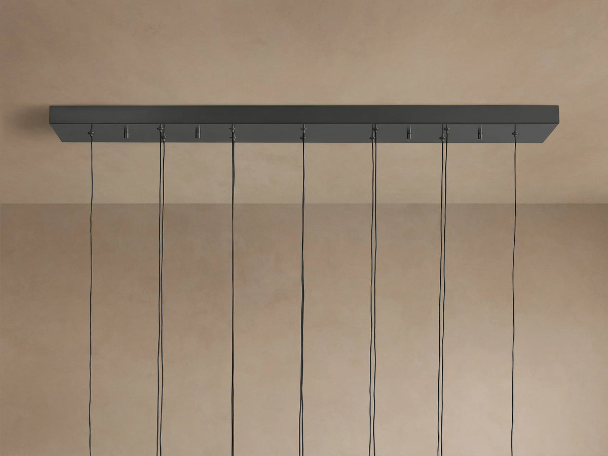 Soltaire Linear Chandelier showing a dark gray linear ceiling mount with multiple black cords hanging down against a light brown wall.