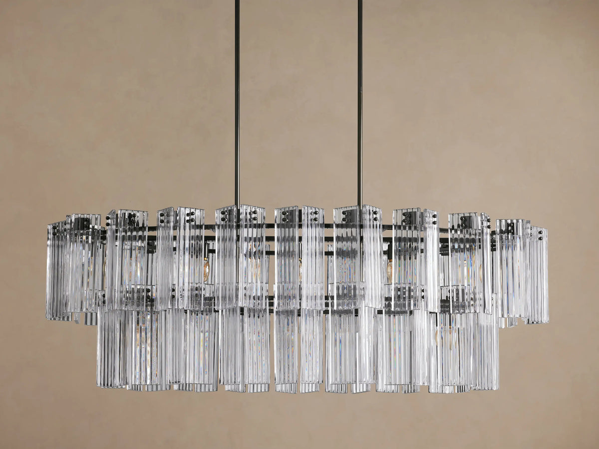 Delsie Oval Tiered Chandelier with two rows of clear ribbed glass rods on a dark metal frame, glowing, against a brown wall.