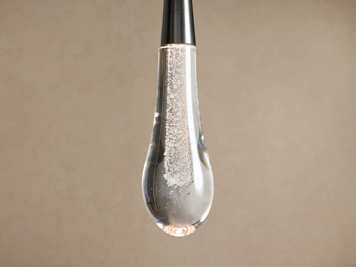 Nickel Soltaire Pendant with a clear, bubbly teardrop glass shade, hanging vertically against a light brown wall.