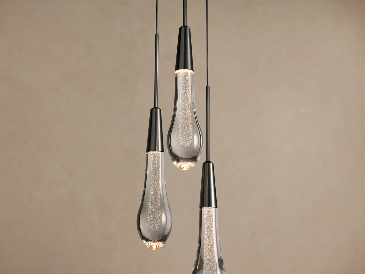 Three Soltaire Cluster Pendants with dark caps & clear, bubble-filled tear-drop glass shades, hanging against a beige wall.