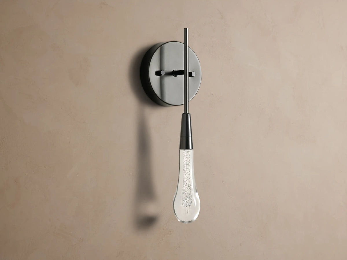 Soltaire Sconce: Dark circular backplate on a taupe wall with a vertical rod holding an illuminated, textured glass teardrop light.