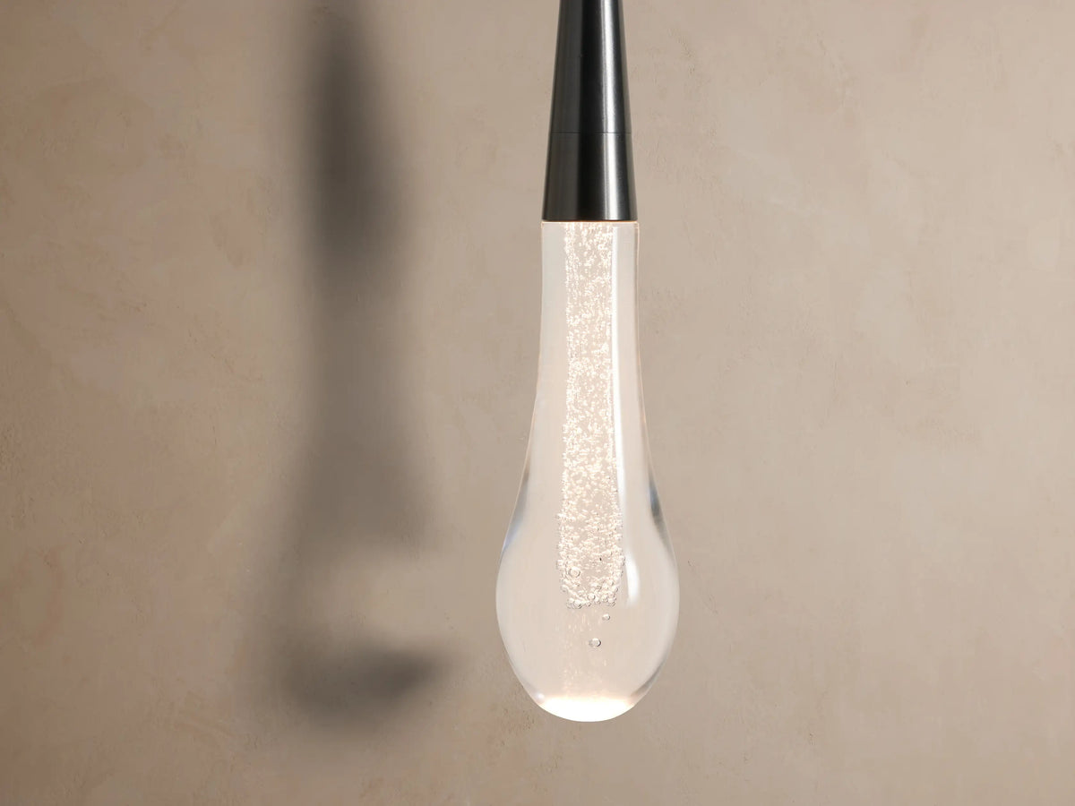 Soltaire Sconce: Dark bronze circular backplate on a tan wall, with a slender rod holding a clear bubbled glass teardrop.