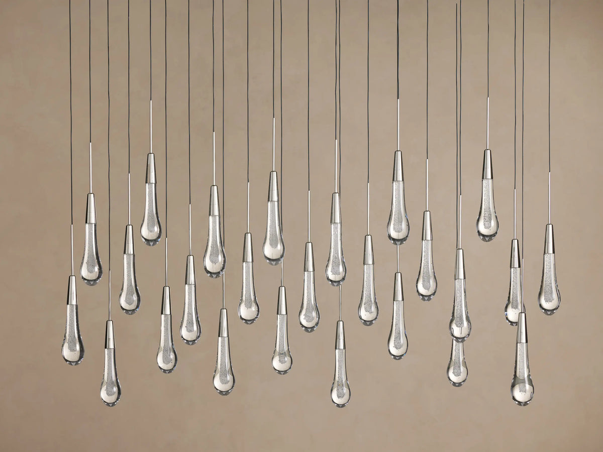Clear Soltaire Linear Chandelier with numerous illuminated teardrop glass pendants, silver caps, and dark cords against a brown background.