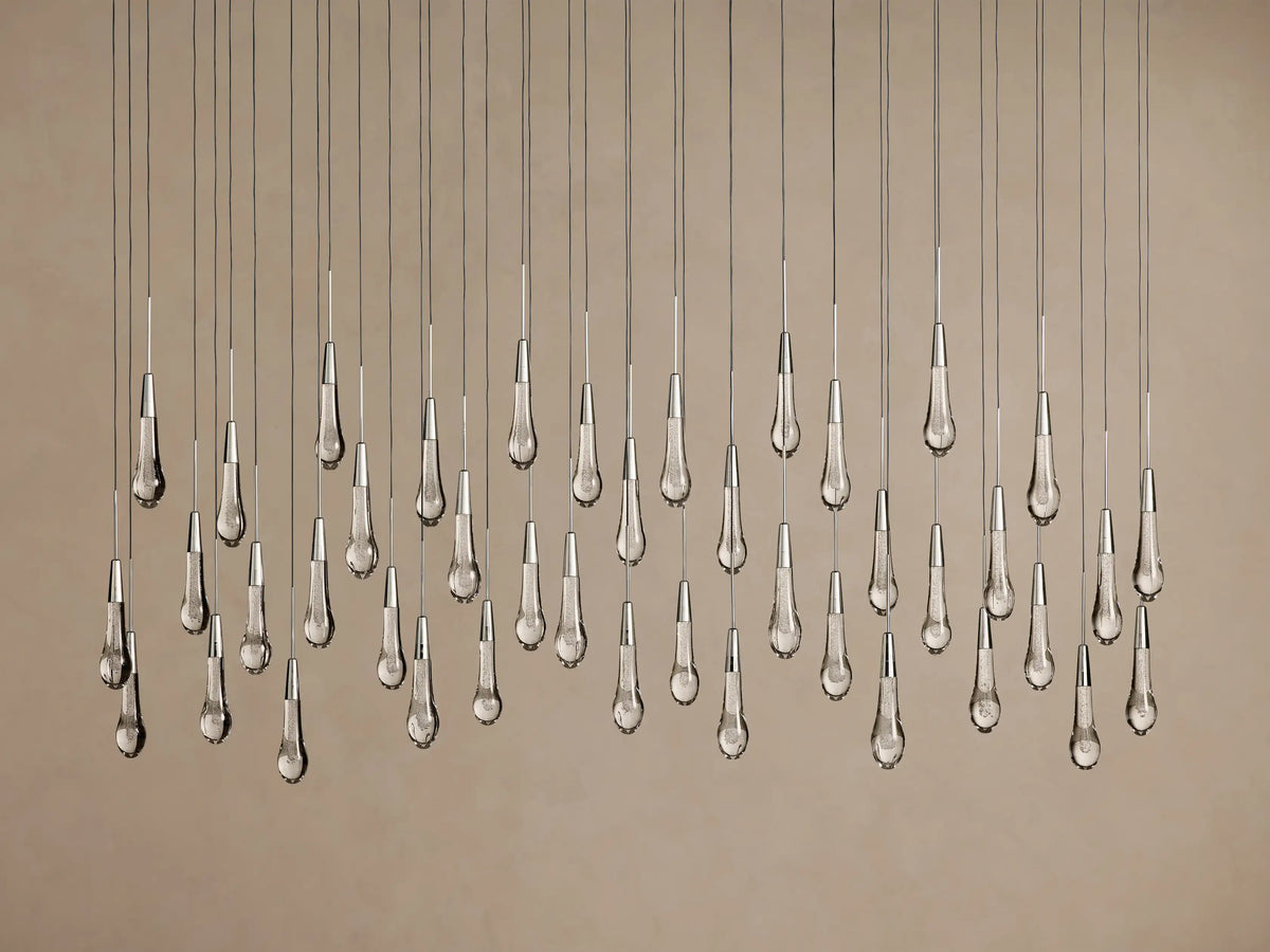 Soltaire Linear Chandelier: clear, textured teardrop glass pendants with silver tops hang illuminated at varying heights on a neutral background.