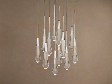 Soltaire Round Chandelier: Clear glass teardrop pendants with metallic caps, glowing, hanging at varied heights on brown background.