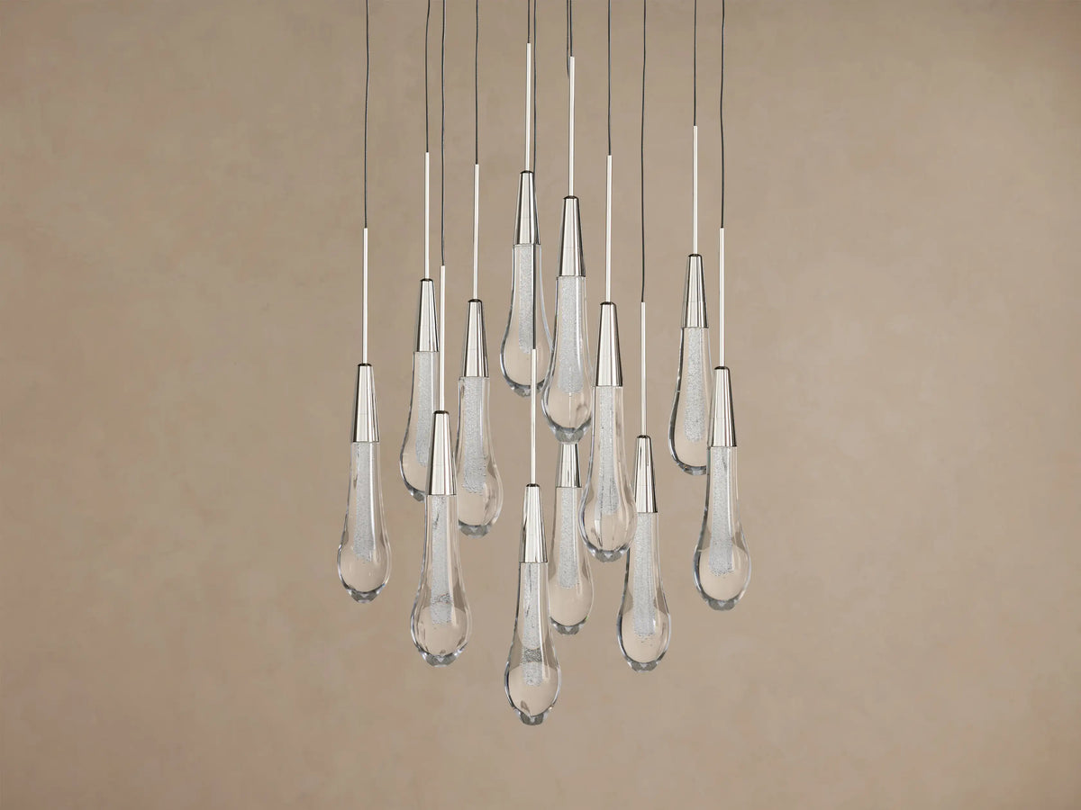 Soltaire Round Chandelier: Clear glass teardrop pendants with metallic caps, glowing, hanging at varied heights on brown background.