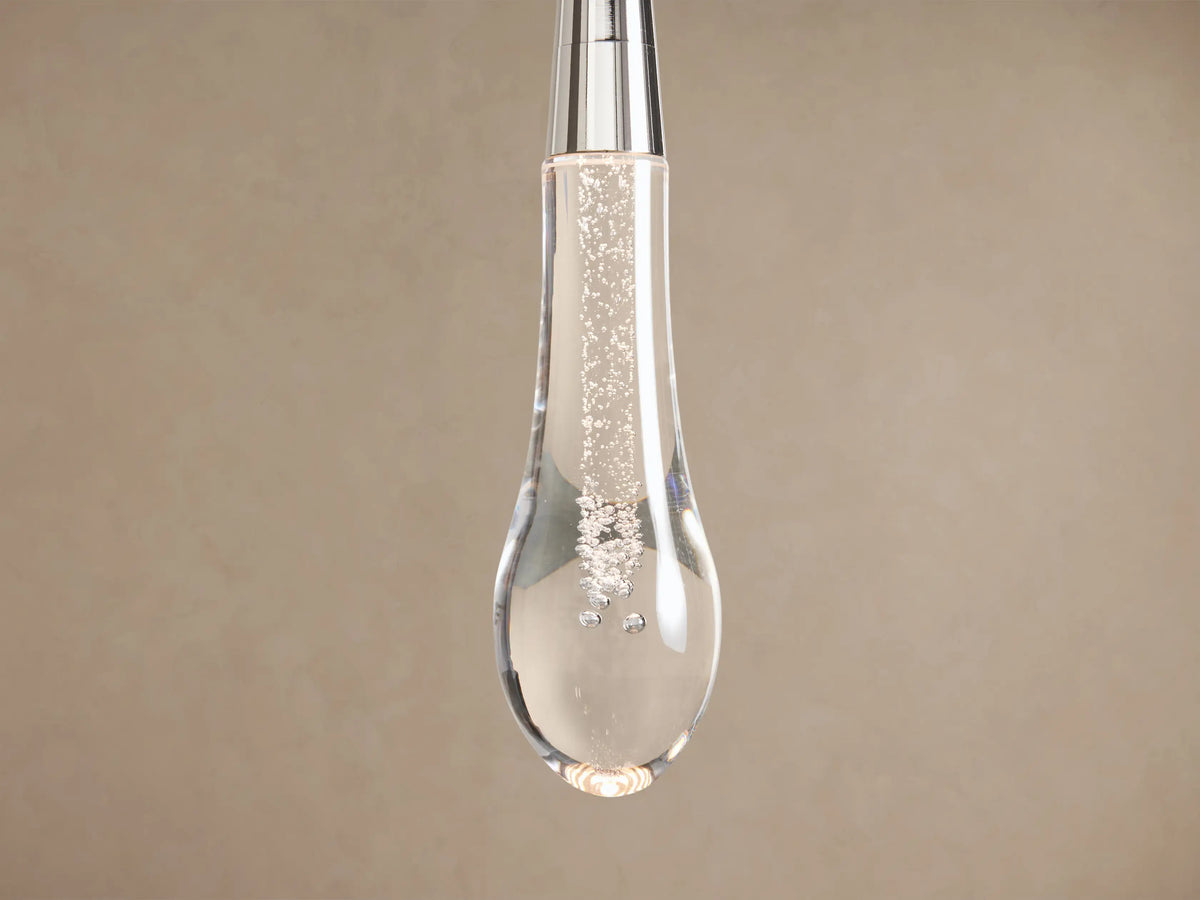 Soltaire Pendant: clear glass teardrop fixture with internal bubbles, silver top & stem, hanging vertically against a beige wall.