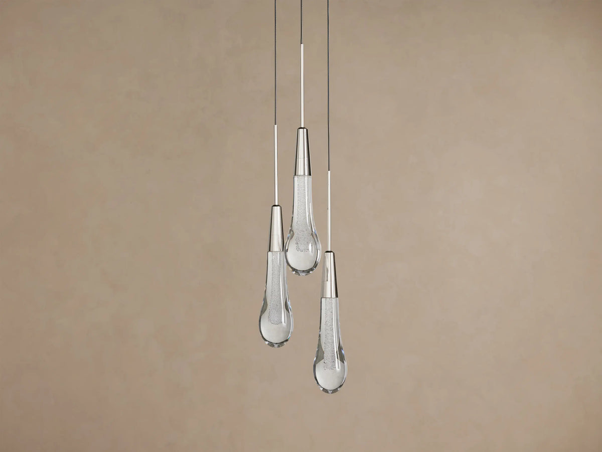 Three Soltaire Cluster Pendants with clear tear-drop glass shades, silver caps, and glowing textured interiors hanging against a brown wall.