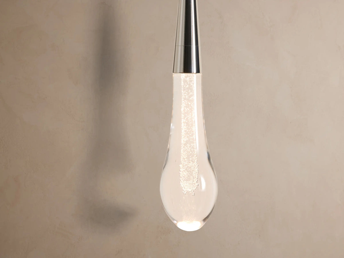 Soltaire Sconce: Round polished nickel backplate with a vertical rod suspending a clear teardrop glass pendant on a light wall.