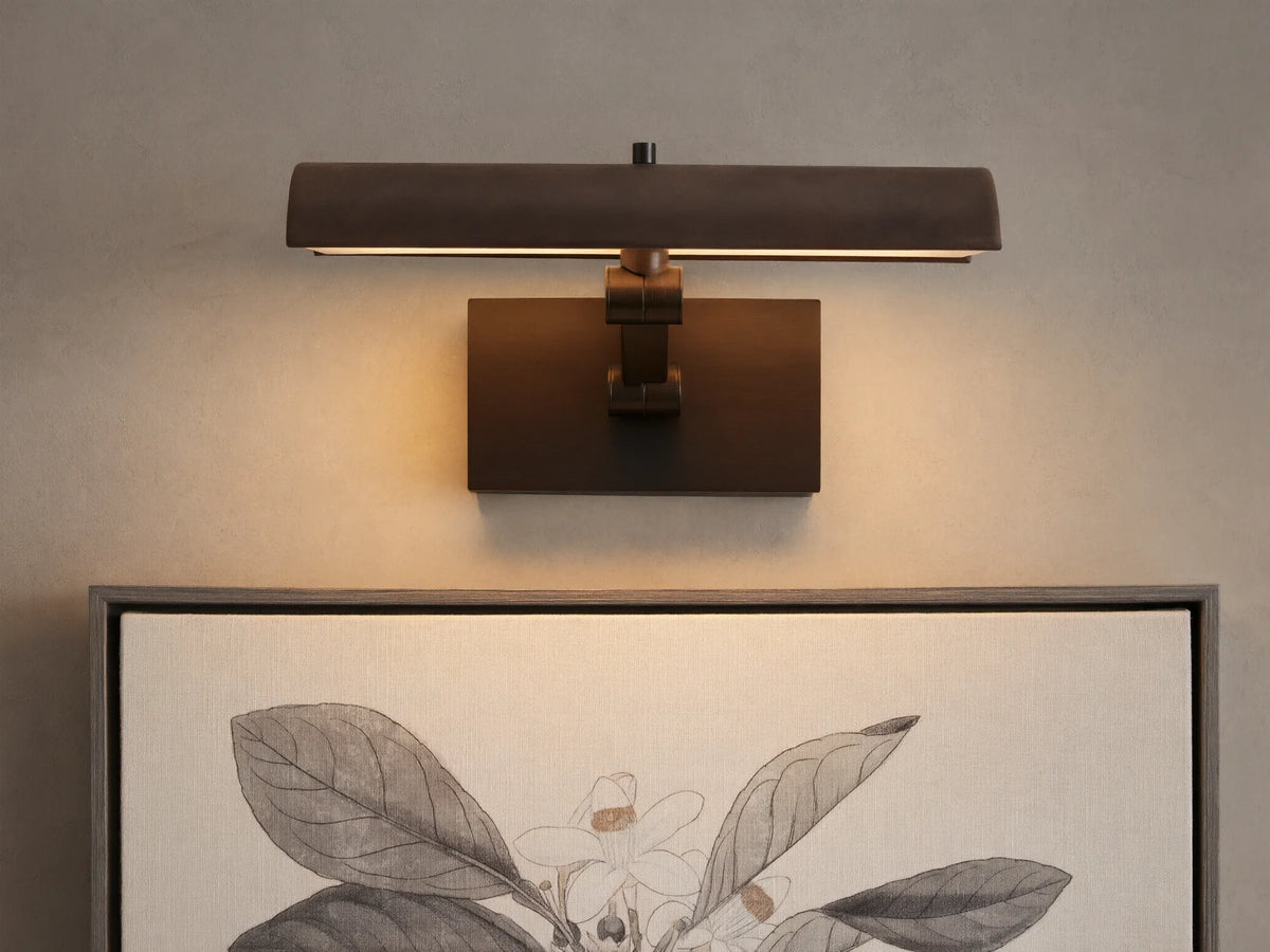 Abel Picture Light Sconce, a dark bronze curved light bar with warm LED light, mounted on a wall above a framed botanical artwork.