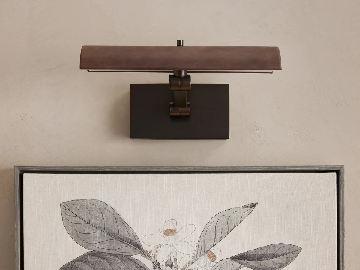 Abel Picture Light Sconce, a dark bronze curved light bar with warm LED light, mounted on a wall above a framed botanical artwork.