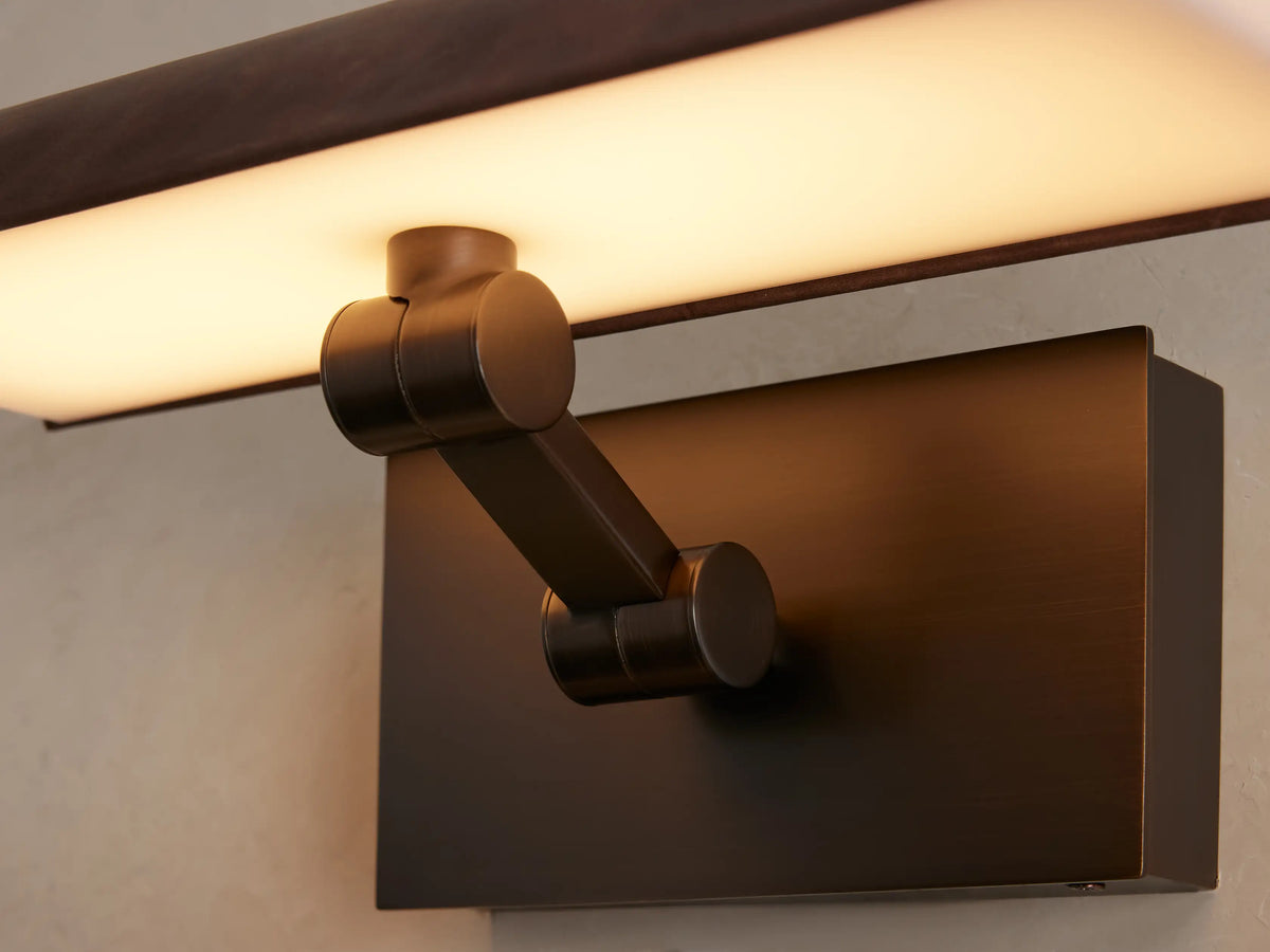 Close-up of Abel Picture Light Sconce. Dark leather-wrapped housing emits warm LED light onto a textured light wall. Brushed bronze backplate.
