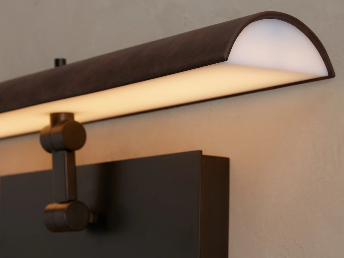 Abel Picture Light Sconce close-up: dark brown leather-wrapped bar with warm LED glow, connecting to a brushed bronze arm & backplate.