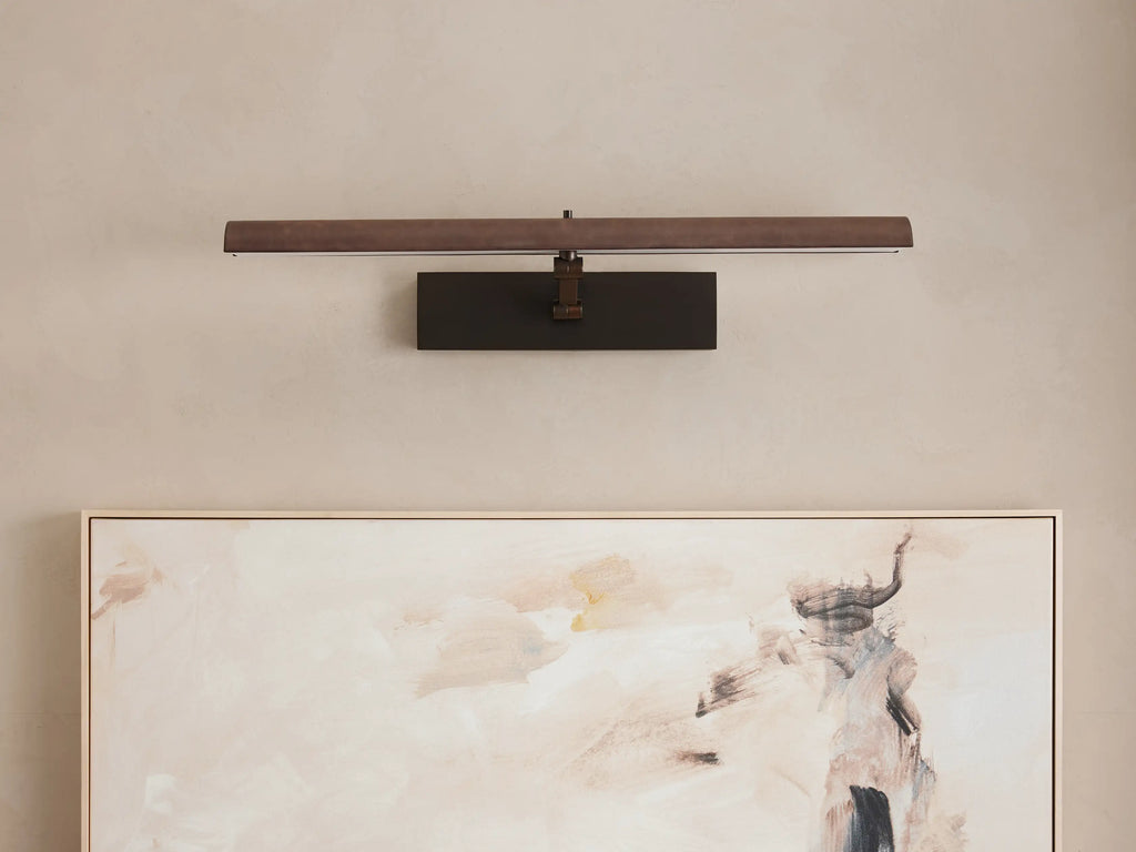 Abel Picture Light Sconce with a dark bronze finish, mounted horizontally on a light wall, casting a warm glow onto an abstract painting.