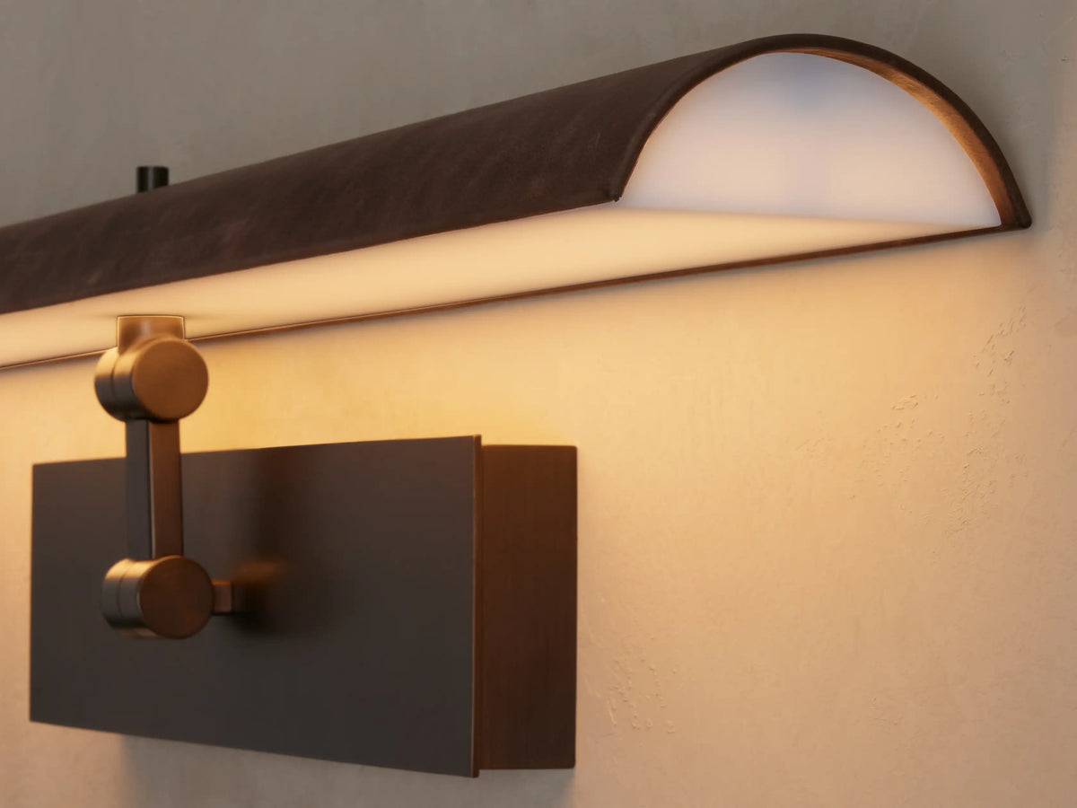 Horizontal Abel Picture Light Sconce with dark leather-wrapped bar emitting warm LED light, mounted on a brushed bronze backplate.