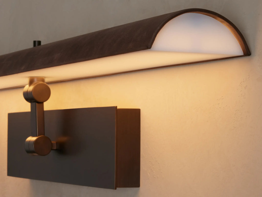 Horizontal Abel Picture Light Sconce with dark leather-wrapped bar emitting warm LED light, mounted on a brushed bronze backplate.
