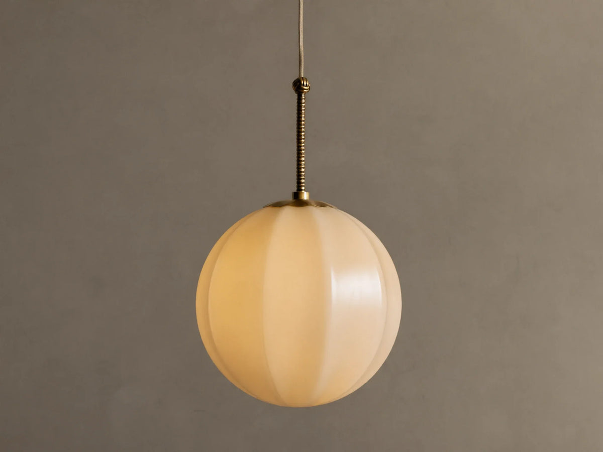 Agnes Pendant: Spherical cream paneled glass shade illuminated, hanging from ribbed antique brass rod against a gray wall.