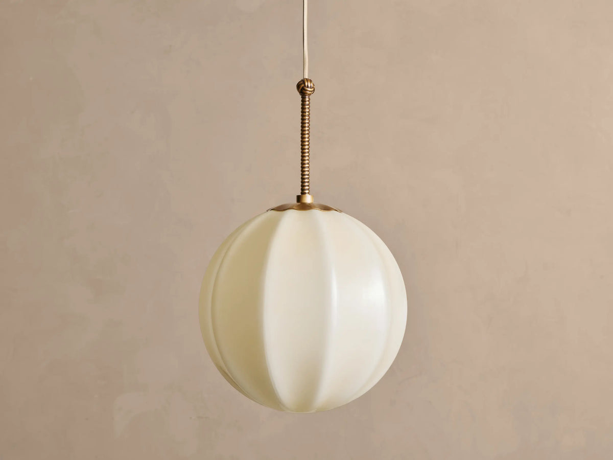 Agnes Pendant: Spherical cream paneled glass shade illuminated, hanging from ribbed antique brass rod against a gray wall.