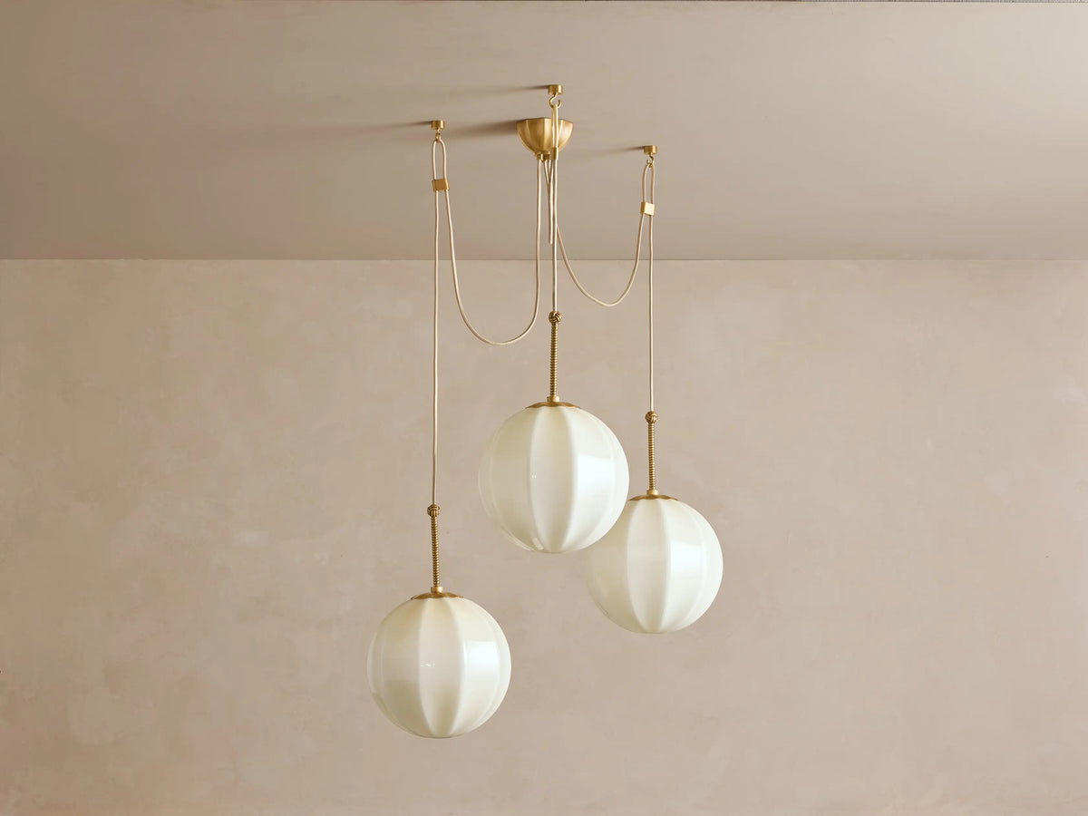 Agnes Chandelier: 3 glowing, paneled cream glass globes suspended from antique brass hardware, draped on a light ceiling.