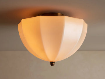 Agnes Flush Mount with paneled cream glass shade & antique brass details, emitting a warm glow from a ceiling mount.