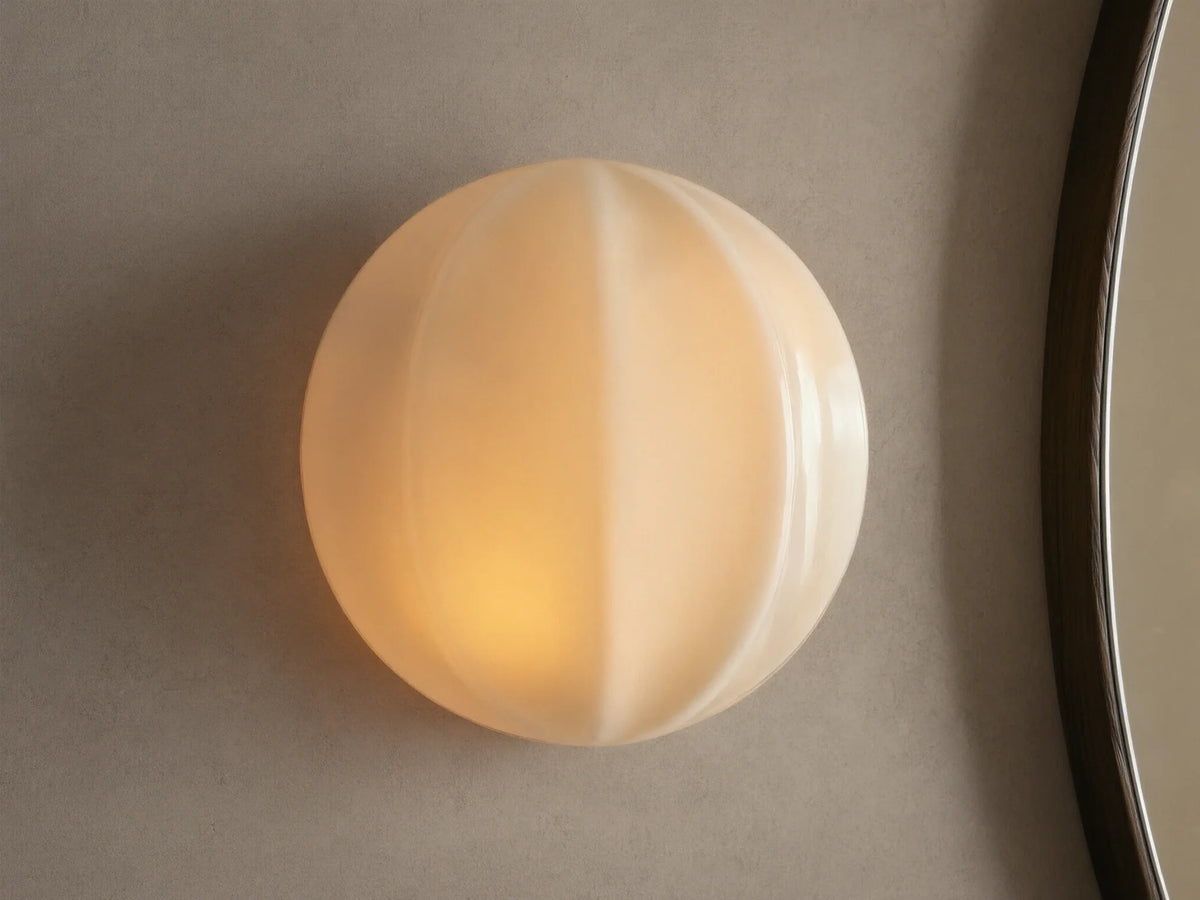 Agnes Clamshell Sconce: Spherical cream paneled glass sconce, glowing warmly, mounted on a light wall next to a dark mirror.